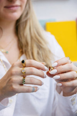 A close-up of a woman's hands wearing multiple gold rings, including one with a black oval stone and another with green stones, while wearing a white blouse.